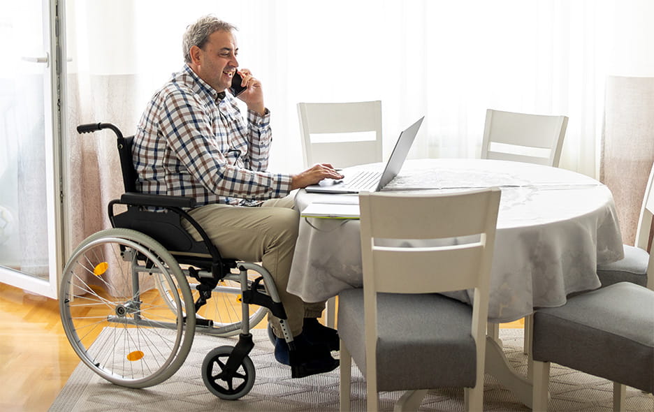 a man in a wheelchair sitting at a table while on the phone and using his computer