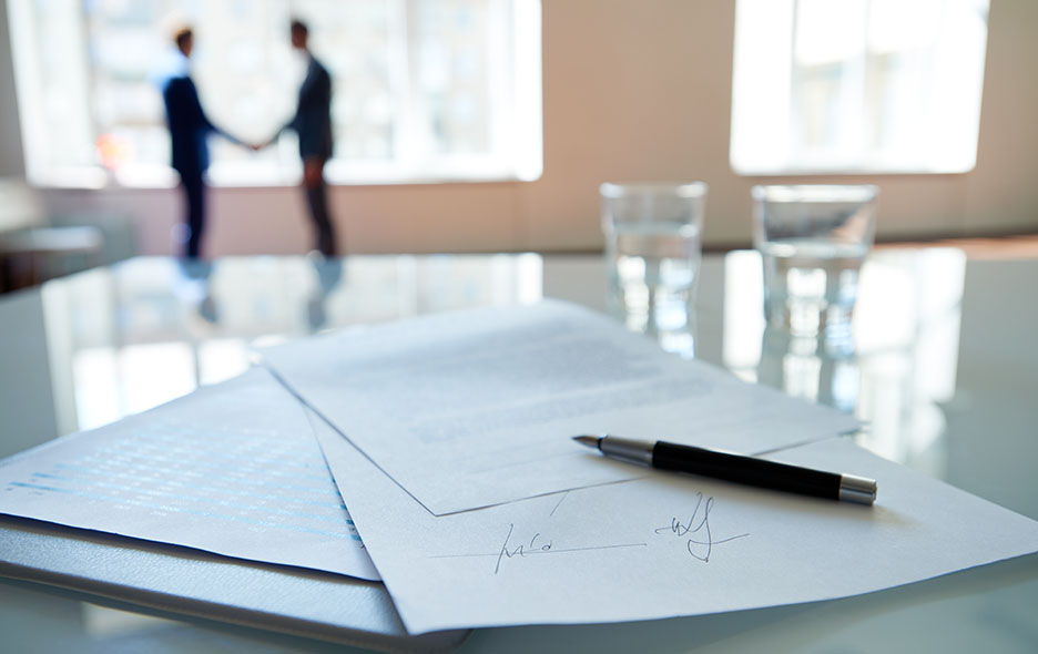 A close-up photo of a business contract and business people shaking hands in the background